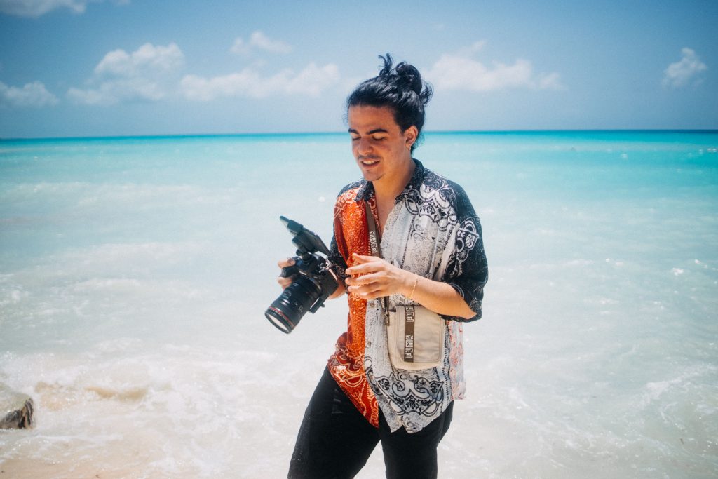 A photographer with a camera walks along a sunny beach by the ocean.